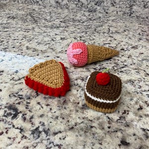 May include: A collection of crocheted food items: a pie slice, an ice cream cone, and a small cake. The pie has a tan crust and red filling. The ice cream cone has a pink scoop. The cake is brown with a white stripe and a red cherry.