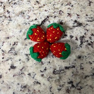 May include: Four crocheted strawberries in a circular arrangement. Each strawberry is red with yellow seed detailing and a green leafy top. They are placed on a speckled, light-coloured surface.