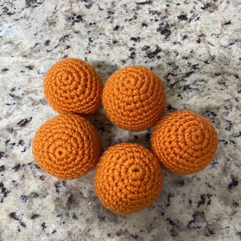 May include: Five handmade orange crochet balls arranged on a speckled surface. Each ball is round and features a spiral pattern at the centre. The balls are a vibrant, solid orange colour.