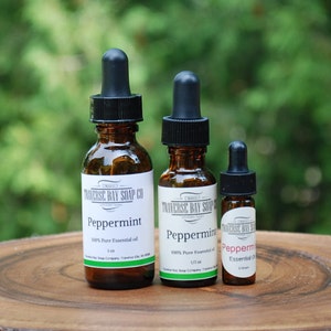 May include: Three amber glass bottles with black dropper tops containing peppermint essential oil. The bottles are labeled "Peppermint 100% Pure Essential Oil" and are arranged in order of size from left to right on a wooden surface.