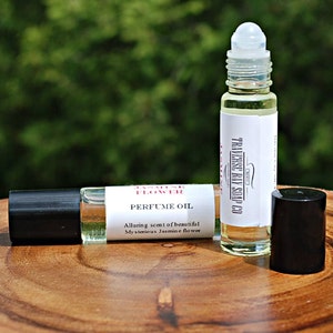 May include: Two clear glass roller bottles with black caps and white labels. The labels read "Jasmine Flower Perfume Oil" and "The Essence Oil Co.". The bottles are sitting on a wooden surface.