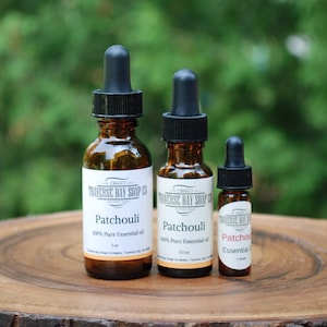 May include: Three amber glass bottles with black dropper tops containing Patchouli essential oil. The bottles are labeled "Traverse Bay Soap Co." and are arranged in order of size from left to right on a wooden surface.