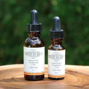 May include: Two amber glass bottles with droppers, labeled "Traverse Bay Soap Co.", containing natural vitamin E tocopherols T-50, 100% pure. The bottles are on a wooden surface.
