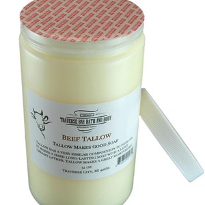 May include: A cylindrical container of off-white beef tallow with a red and white tamper-evident seal on the lid. The white label features a cow illustration, the text Traverse Bay Bath and Body, Beef Tallow, and 32 oz, with a detached white lid.