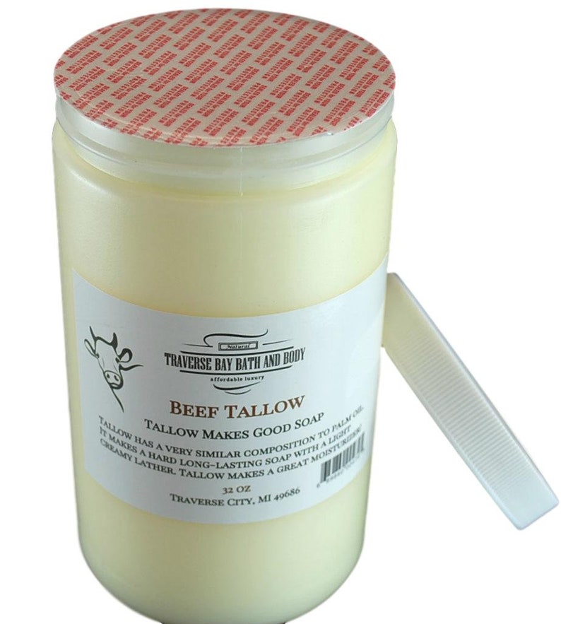 May include: A cylindrical container of off-white beef tallow with a red and white tamper-evident seal on the lid. The white label features a cow illustration, the text Traverse Bay Bath and Body, Beef Tallow, and 32 oz, with a detached white lid.