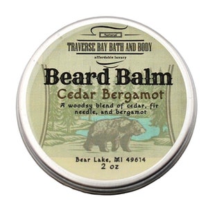May include: A silver tin with a label that reads "Natural Traverse Bay Bath and Body affordable luxury Beard Balm Cedar Bergamot A woodsy blend of cedar, fir needle, and bergamot Bear Lake, MI 49614 2 oz". The label features a cartoon illustration of a brown bear walking through a forest.