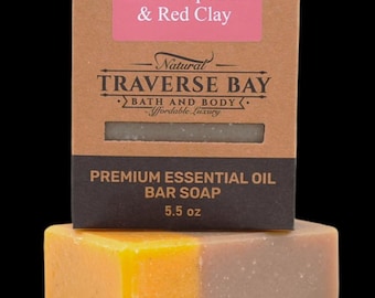 May include: A rectangular bar of soap with a yellow and light brown color gradient, positioned in front of a brown cardboard box. The box features the text Pink Grapefruit & Red Clay, Natural TRAVERSE BAY BATH AND BODY, and Premium Essential Oil Bar Soap 5.5 oz.