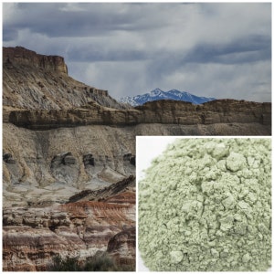 May include: A close-up of a pile of green clay powder. The powder is a light green color and has a rough texture. The powder is in front of a background of a rocky landscape with a mountain range in the distance.