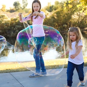 May include: Two young girls are playing with a large, iridescent bubble outdoors. One girl, wearing a pink shirt and jeans, is holding the bubble wand. The other girl, in a white shirt and blue pants, is watching the bubble float.