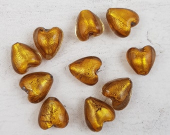 May include: Ten gold heart-shaped glass beads with a metallic finish. The beads are scattered on a white surface.