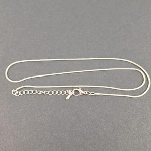 May include: A silver snake chain necklace with an adjustable length. The necklace features a delicate, thin chain and a lobster clasp. The necklace is shown on a grey surface.