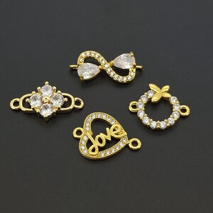May include: Four gold-toned jewellery connectors featuring clear cubic zirconia stones. The set includes an infinity symbol, a four-petaled flower shape, a circular wreath with a small bow, and a heart-shaped charm with the word love in script.