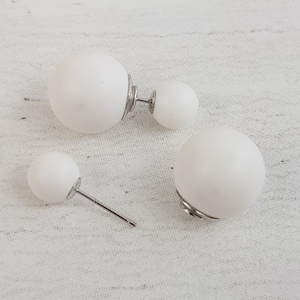 May include: A pair of white double-sided ball earrings with silver posts. The earrings are on a white fabric background.