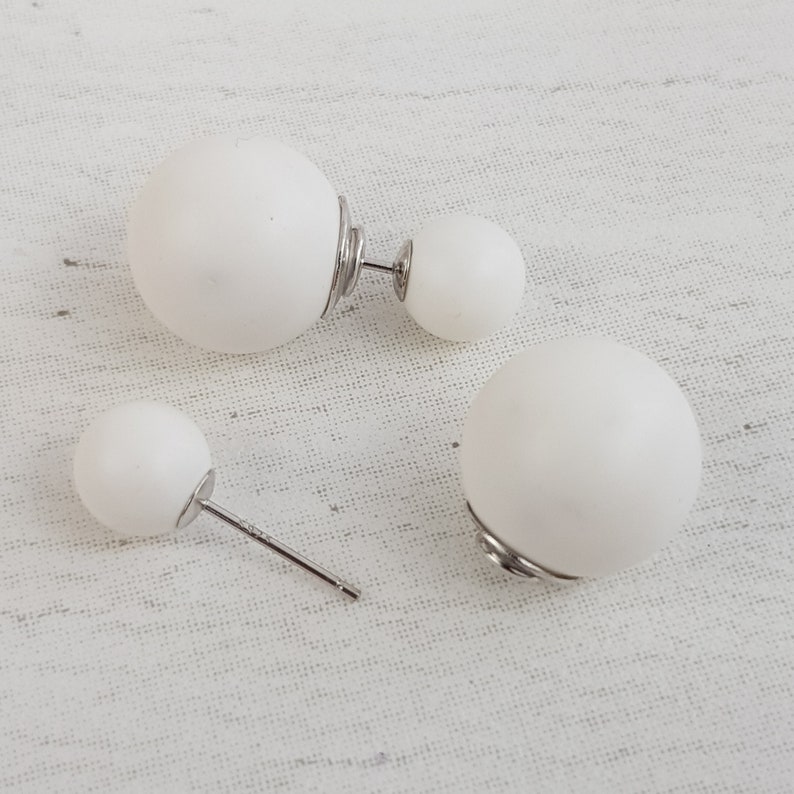 May include: A pair of white double-sided ball earrings with silver posts. The earrings are on a white fabric background.