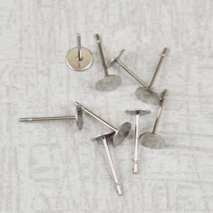 Può includere: Una collezione di perni per orecchini color argento con cuscinetti piatti e rotondi. I perni sono dritti con un piccolo cuscinetto rotondo per attaccare l'orecchino. Gli orecchini sono sparsi su una superficie grigio chiaro.