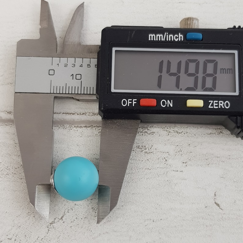 May include: A turquoise blue bead is measured with a digital caliper. The caliper reads 14.98 mm.