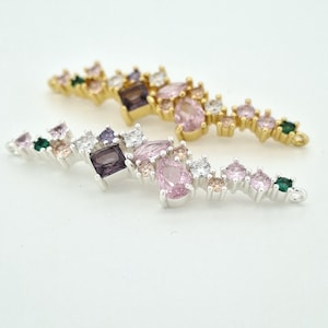 Pode incluir: Dois conectores de joias alongados com grupos de pedras preciosas de várias formas em tons de rosa, roxo, transparente e verde. Um conector é montado em metal dourado e o outro em metal prateado, ambos com argolas em ambas as extremidades.
