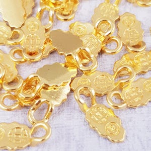 May include: A close-up of a pile of gold-colored metal jewelry findings. The findings are shaped like a small oval with a loop on top. The findings are all identical and have a textured surface.