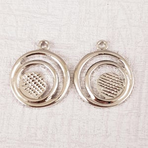 May include: A pair of silver-toned metal earring blanks with a circle design. Each blank has a textured circle in the center and a loop at the top for attaching a finding.