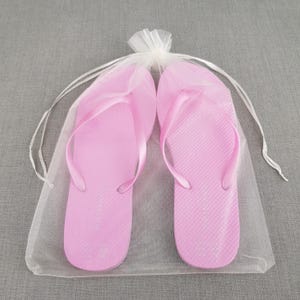 May include: Two pairs of pink flip flops in a white tulle bag. The flip flops have a simple design and are perfect for a beach holiday or a summer wedding.