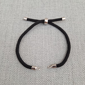 以下が含まれることがあります： 銀色の金属製の留め具と銀色のビーズのアクセントが付いた、黒い編み紐のブレスレットです。