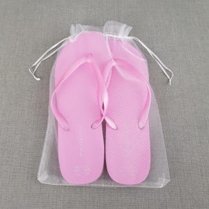 Op de afbeelding: Twee roze slippers in een witte mesh tas. De slippers hebben het woord "PINK" op de bandjes gedrukt.