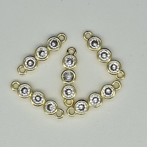 May include: Three gold-toned jewelry connectors, each featuring a vertical row of four clear, round, bezel-set stones. Each connector has a small circular loop at both the top and bottom ends, designed for linking components in jewelry making projects.