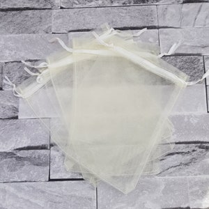 Op de afbeelding: Een stapel doorschijnende organza-geschenkzakjes in ivoor met witte trekkoordjes. De zakjes zijn gemaakt van een delicate, doorschijnende stof en zijn perfect voor het verpakken van kleine cadeautjes of attenties.