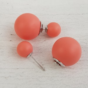 May include: A set of three coral-colored double-sided stud earrings. The earrings are made of a smooth, matte material and have a simple, modern design.