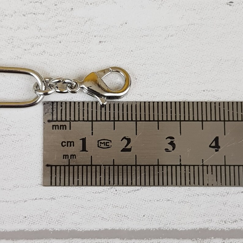 Może przedstawiać: Zbliżenie srebrnego łańcuszka z karabińczykiem. Łańcuszek jest mierzony linijką, pokazując, że ma około 2 cm długości.