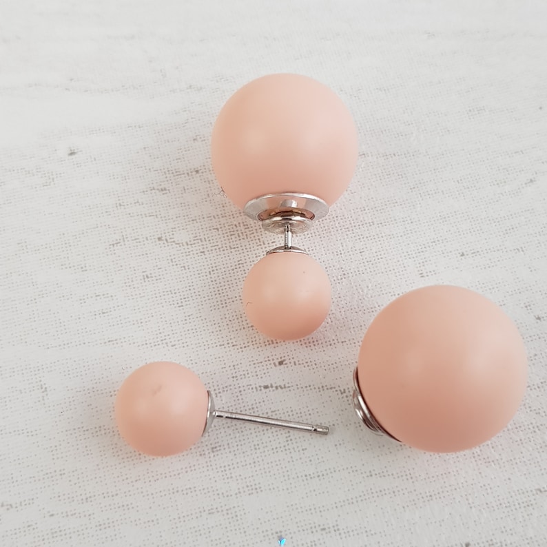 May include: A pair of peach-colored double-sided stud earrings. The earrings have a larger ball on one side and a smaller ball on the other side.
