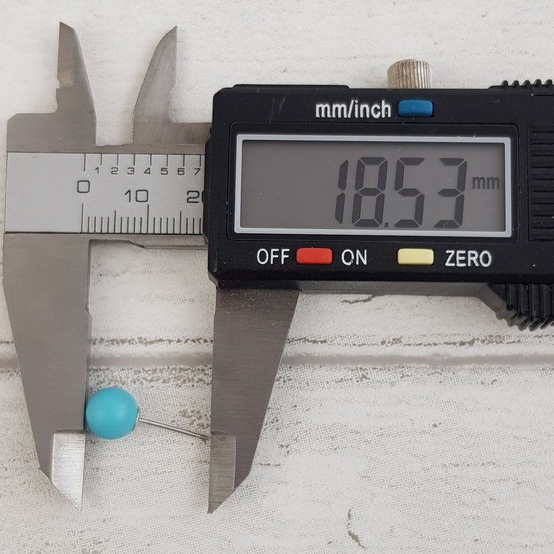 May include: A blue bead with a silver pin is measured with a digital caliper. The caliper reads 18.53 mm.