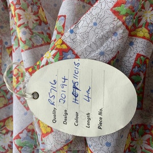 May include: A white fabric with a red, yellow, and blue floral pattern. A white tag with black writing is attached to the fabric. The tag reads "Quality: RS716, Design: 20194, Colour: H 6785/11(15), Length: 4m, Piece No."