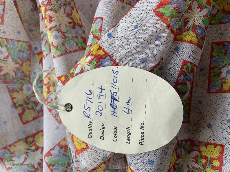 May include: A white fabric with a red, yellow, and blue floral pattern. A white tag with black writing is attached to the fabric. The tag reads "Quality: RS716, Design: 20194, Colour: H 6785/11(15), Length: 4m, Piece No."
