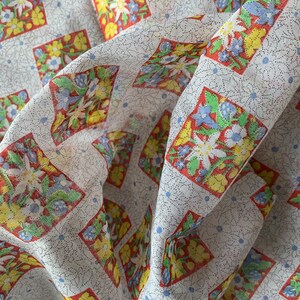 May include: White fabric with a repeating pattern of red, yellow, and blue floral squares. The fabric is folded and has a textured surface.