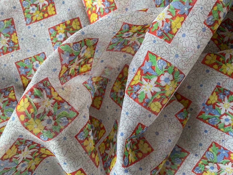 May include: White fabric with a repeating pattern of red, yellow, and blue floral squares. The fabric is folded and has a textured surface.