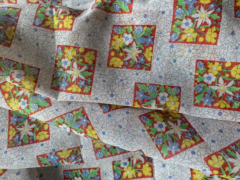 May include: White fabric with a repeating pattern of red, yellow, and blue floral designs. The floral designs are in a diamond shape and are surrounded by a white background with small blue dots.