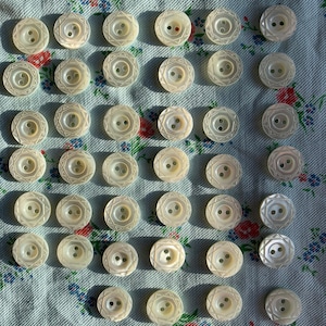 May include: A collection of 40 white mother-of-pearl buttons with a geometric design. The buttons are arranged in a heart shape on a light blue fabric with floral patterns.