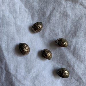 May include: Five small, metallic buttons with a textured, irregular surface that resembles the appearance of a walnut shell. Each button has a rounded, dome-like shape with a dark, bronze-toned finish and a reflective, uneven surface texture.