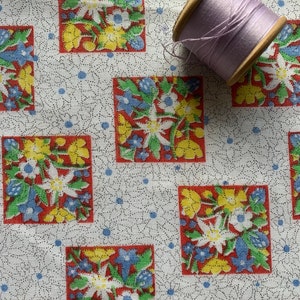 May include: White fabric with a repeating pattern of red, yellow, and blue floral squares. A spool of lavender thread is on top of the fabric.