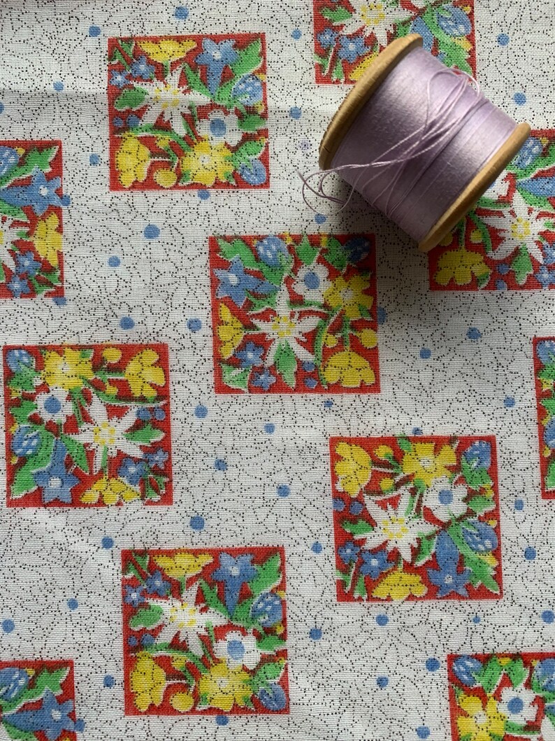 May include: White fabric with a repeating pattern of red, yellow, and blue floral squares. A spool of lavender thread is on top of the fabric.