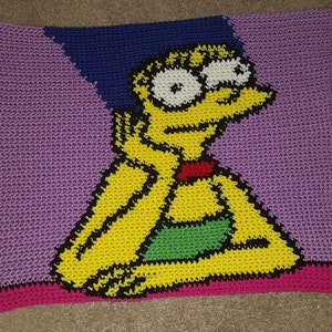 Op de afbeelding: Gehaakte deken met een geel cartoonfiguur met blauw haar, een rode strik en groene kleding. De deken is gemaakt van paars, blauw, groen, geel, zwart en roze garen.