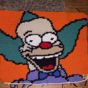 Op de afbeelding: Een gehaakt kussen met een cartoonfiguur met een brede glimlach, een rode neus en groen haar. Het personage draagt een paarse en blauwe outfit met een zwarte strik. Het kussen is geplaatst tegen een oranje achtergrond.