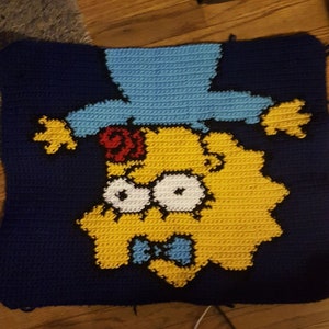 Op de afbeelding: Een vierkant kussen van gehaakt garen met een geel cartoonfiguur met blauwe ogen, een rode bloem en een blauwe hoed. Het figuur draagt een blauwe strik en heeft een gele en zwarte omtrek.