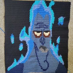 May include: A handmade crochet piece featuring a character with blue flames and a dark background. The character's face is detailed with brown eyes, a gray nose, and a blue outline. The piece is square-shaped and appears to be a blanket or wall hanging.