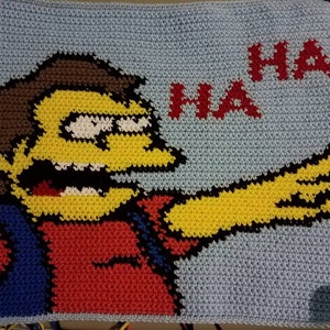 Op de afbeelding: Een gehaakt deken met een geel cartoonfiguur met bruin haar, een rood shirt en een blauwe broek. Het personage zegt "HA HA" in rode letters.
