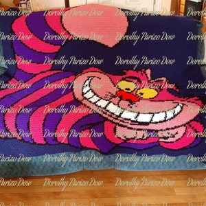 Puede incluir: Una manta de ganchillo con un gato de Cheshire sonriente de Alicia en el País de las Maravillas. El gato es rosa con ojos amarillos y dientes blancos. La manta es una mezcla de morado, rosa y azul.