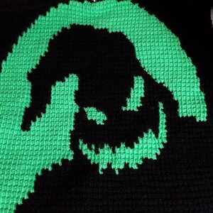 Op de afbeelding: Een vierkant van zwart en groen gehaakt garen met een silhouet van een grijnzend gezicht.
