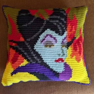 Puede incluir: Una almohada de ganchillo con un retrato colorido de Maléfica de La Bella Durmiente de Disney. La almohada está hecha con una variedad de colores, incluyendo morado, azul, verde, amarillo y rojo. La almohada es perfecta para agregar un toque de magia de Disney a cualquier habitación.