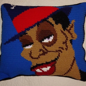 May include: A crocheted pillow featuring a cartoon character with a red hat, brown skin, and a large, toothy grin. The background is blue.